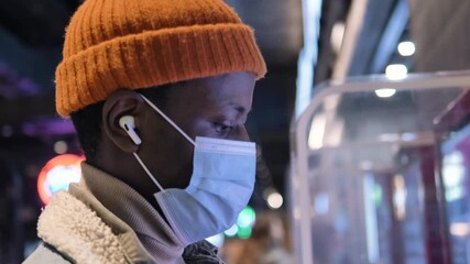 Wall Mural - Black guy consumer in orange hat and disposable mask with wireless headphones looks into interactive display choosing burgers closeup