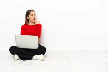 Young caucasian woman with a laptop sitting on the floor yawning and covering wide open mouth with hand