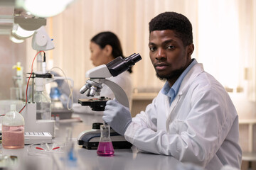 Sticker - Young African male chemist with microscope sitting by desk in laboratory