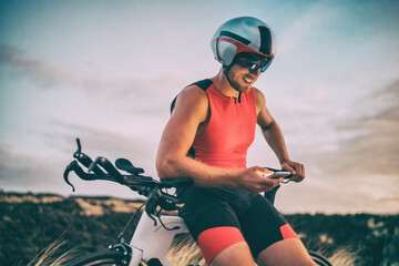 Triathlon cyclist athlete man using health monitor sports app on mobile phone after bike training. Triathlete at dusk wearing tri suit and helmet on technology device.