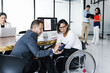 © Marcos - Latina transgender businesswoman in wheelchair with office colleague checking email on smartphone and office employees working in the background, in disability concept and disabled people