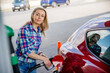 Pretty blonde woman is filling her red car with gasoline at a gas station.