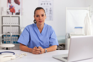 Wall Mural - Portrait of pretty medical nurse smiling at camera in hospital office wearing blue uniform. Healthcare practitioner sitting at desk using computer in modern clinic looking at monitor, medicine.