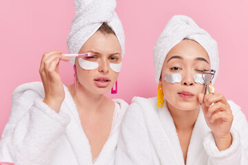 Photo of serious attentive young women do professional make up use cosmetic brush eyelashes curler apply collagen patches under eyes dressed in white bathrobes isolated over pink background.