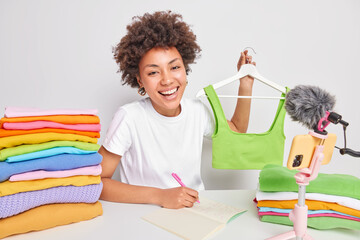 Smiling female fashion blogger demonstrates trendy clothes sells green top online makes notes about customer orders poses in front of camera at white table with colorful folded laundry near.