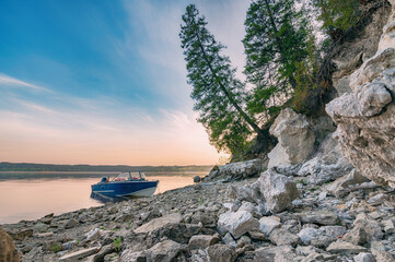 Wall Mural - An evening landscape, a boat on a steep, rocky riverbank at sunset, a sloping fir tree on a cliff, a pink blue sky.