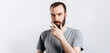 © Daria Lukoiko - Serious puzzled handsome male model with beard holding hand on chin as if thinking about something, squinting at camera with suspicious look and standing over gray background. Man decides what to buy