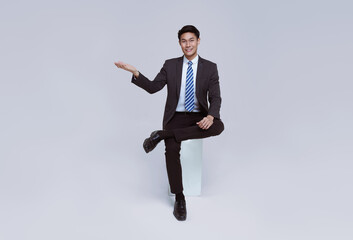 Handsome and friendly face asian businessman smile in formal suit sitting on chair points his hands to presented on white background studio shot.