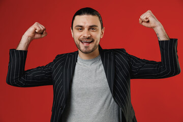 Wall Mural - Young strong sporty businessman latin man in black striped jacket grey shirt show biceps muscles on hand demonstrating strength power isolated on red background studio. Concept of people lifestyle