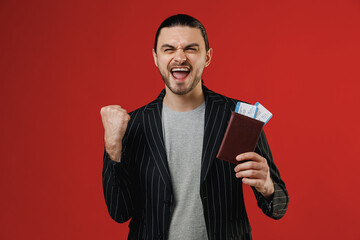 Traveler tourist latin man in black jacket grey shirt hold passport ticket do winner gesture clench fist isolated on red background. Passenger travel abroad weekend getaway Air flight journey concept