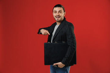 Wall Mural - Young smiling successful businessman latin man 20s with ponytail wearing black striped jacket grey shirt point finger on briefcase diplomat looking camera isolated on red background studio portrait