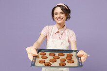Young Satisfied Happy Housewife Housekeeper Chef Cook Baker Woman In Pink Apron Showing Chocolate Cookies Biscuits On Baking Sheet Isolated On Pastel Violet Background Studio. Cooking Food Concept