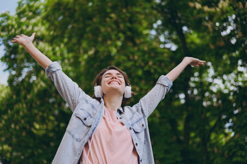 Bottom view young carefree woman in jeans clothes headphones listen to music walk stroll in green park with raised up hands outdoors with green blur trees on background People urban lifestyle concept