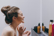 © ViDi Studio - Side view fun half naked topless young woman 20s applying moisturizer, anti-aging facial cream stand in front of mirror in bathroom do morning routine Skin care healthcare cosmetic procedures concept.