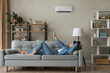 © fizkes - Full length relaxed young happy woman homeowner lying on comfortable sofa turning on air conditioner with remote controller, breathing fresh air, enjoying lazy weekend time, meditating in living room.