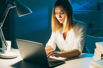 Night work. Remote job. Freelancer lifestyle. Late task. Serious smart workaholic woman using laptop typing at dark workplace with desk lamp light in home office.