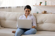 © fizkes - Smiling young mixed race woman sit on couch at home look at laptop screen talk on webcam video digital call. Happy Indian female use computer work online on gadget. Technology, communication concept.
