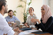 © fizkes - Happy diverse employees, partners discussing startup ideas or project statistics at meeting, smiling Asian muslim businesswoman wearing hijab talking with businessman, having fun, sitting at table