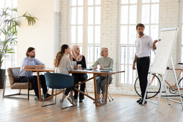 Canvas Print - Businessman coach pointing at diagram on white board, making flip chart presentation to diverse employees, explaining project strategy, training staff, businesspeople listening to mentor at seminar