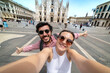 © photomaticstudio - friends couple taking a selfie in front of a famous landmark - Tourists photographing the Duomo cathedral in Milan - happy tourist people on vacation sightseeing the city Milano