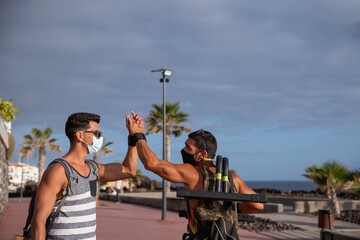 Two male athletes greeting each other, they just arrived at the outdoor training park, they wear masks to train safely during the coronavirus pandemic, it's summer