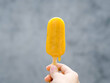 Yellow popsicle in woman hand on grey wall background, melting, summertimes. Female hand holding yellow frozen popsicle ice pop.