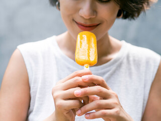 Yellow popsicle in beautiful happy Asian woman’s hand wearing casual white sleeveless shirt, outdoors. Woman eating popsicles. Smiling female enjoying ice lolly in summer.