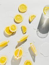 Fresh Lemons Cut Into Slices On Bright White Wet Surface With Glass Of Water In The Corner. Ingredients For Lemonade. Juicy And Ripe Summer Citrus Fruit. Creative Refreshment Drink Concept. Top View.