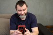 Portrait of adult caucasian man with beard smiling uses phone while sitting on sofa at home