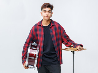 Portrait shot of attractive young teenage drummer standing nearby cymbal in the studio. Young trendy junior musician carrying drumsticks and snare drum while looking to camera with white background