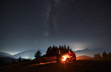 Wall Mural - Young man sitting on mountain hill by campfire playing guitar for his beloved under night starry sky with Milky way. Silhouettes of trees, mountains and light from surrounding villages on the backdrop