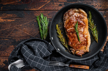 Wall Mural - Grilled new york strip beef meat steak in a pan with herbs. Dark wooden background. Top view. Copy space