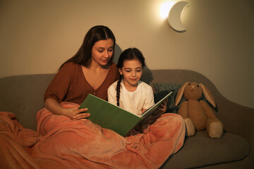 Wall Mural - Little girl with mother reading book in living room lit by night lamp