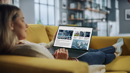 Beautiful Caucasian Female is Using Laptop Computer for Reading Latest News at Home Living Room while Lying on a Couch Sofa. Freelance Specialist Working From Home, Surfing the Internet.