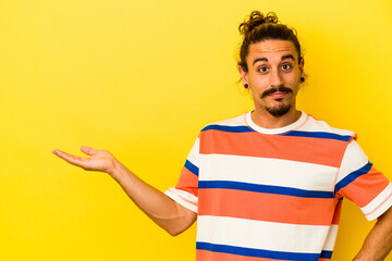 Young caucasian man with long hair isolated on yellow background showing a copy space on a palm and holding another hand on waist.