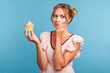© khosrork - Pensive young adult blonde female with funny hair buns holding cake in hand and thinks eat or not, needs to keep diet, looking away with thoughtful look. Indoor studio shot isolated on blue background