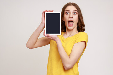 Wall Mural - Young excited woman wearing casual yellow T-shirt, holding digital tablet in hands, showing blank device screen to camera, expressing astonishment. Indoor studio shot isolated on gray background.