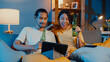 Happy young asian couple enjoy night party event online sit couch use tablet video call with friends toast drink beer via video call online in living room at home, Social distancing concept.