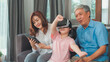 © tirachard - Asian grandparents and granddaughter using virtual reality and tablet play games at home. Senior Chinese, grandpa and grandma happy relax with young girl lying on sofa in living room concept.