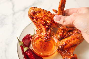 Woman dipping fried chicken wing into bowl with hot honey on light background, closeup