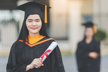 Wall Mural - A young Asian woman university graduate in graduation gown and mortarboard holds a degree certificate stands in front of the university building after participating in college commencement