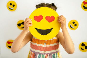 Wall Mural - The girl is holding a happy love emoticon with hearts instead of eyes , covering her face with them. The child's face is hidden behind emojis. World smile day