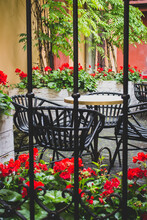 Cozy Street Cafe With Iron Fence In Old Town. Empty Patio With Outdoor Furniture And Red Flowers. Hotel Terrace With Chairs And Tables. Sidewalk Cafe Decoration In Backyard. Beautiful Outdoor Cafe.