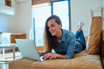 Wall Mural - Casual smart modern smiling millennial girl with wireless headphone using laptop for watching video content, checking social media and shopping online during relax on sofa at home .