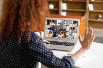 Wall Mural - Close-up of curly woman waving at the laptop screen with people profiles, using an application for distance video communication with coworkers, webinar participants, meeting online in pandemic