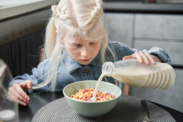 Wall Mural - Girl pouring milk from the bottle to the plate while sitting at the kitchen