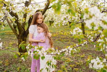 Two Girls Sisters Have Fun And Cuddle In The Garden With Flowering Trees.