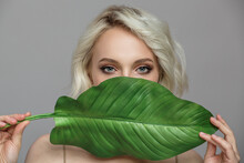 Portrait Of Blonde Woman And Green Leaf. Organic Cosmetics Concept. Gray Background.