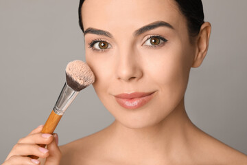 Beautiful young woman applying face powder with brush on grey background, closeup