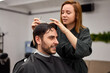 © Andrii - Handsome blue eyed man sitting in barber shop. Hairstylist Hairdresser Woman cutting his hair. Female barber.